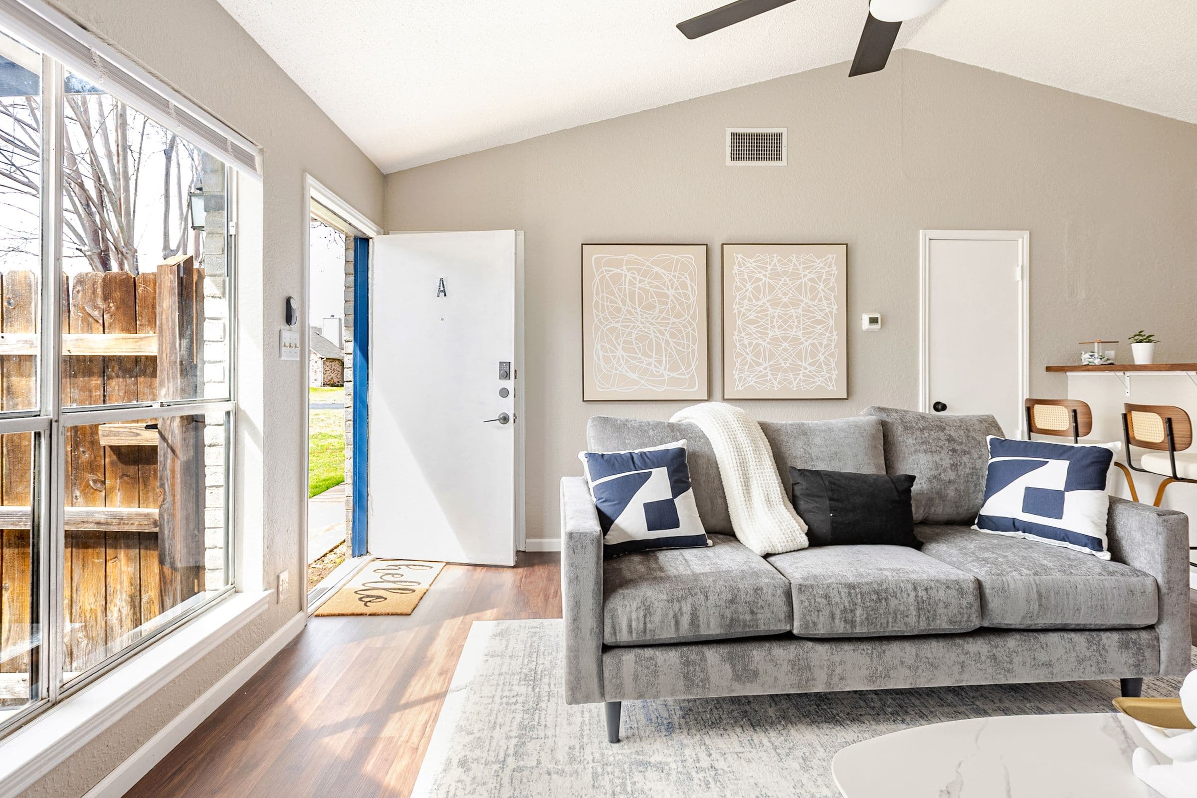 Living room with a grey sofa under a vaulted ceiling, the front door open onto the yard